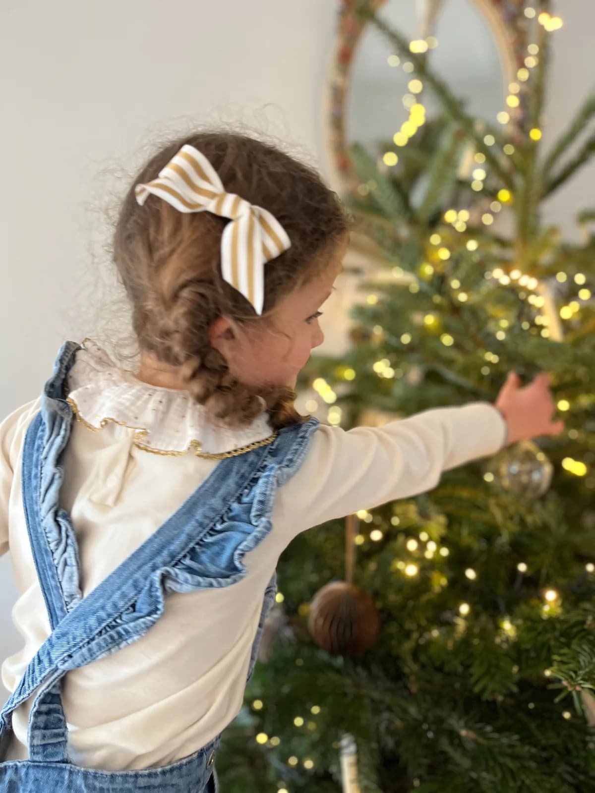 Petite fille décorant le sapin de Noël, blouse blanche fil doré et salopette en jean By Mésange