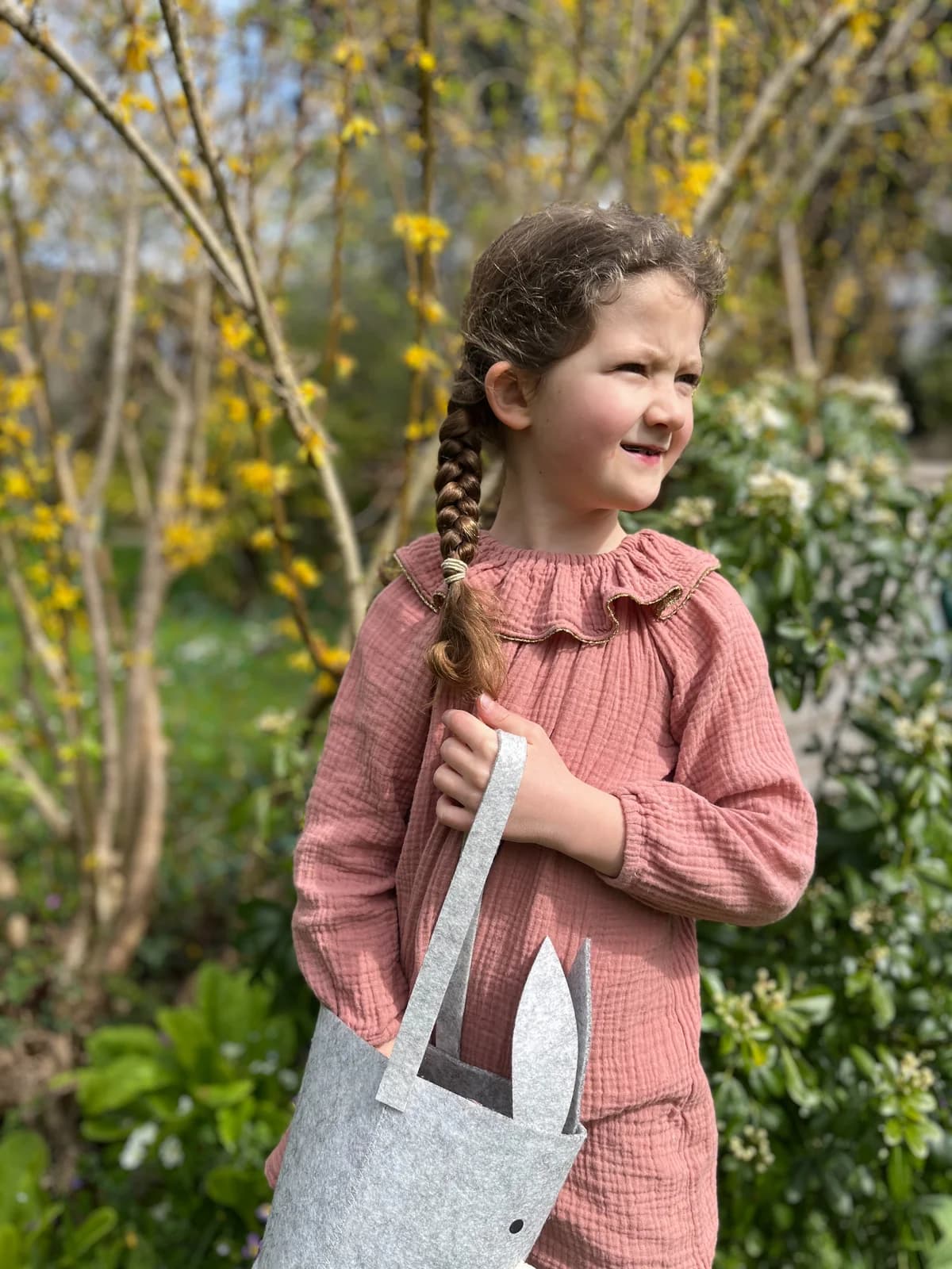Petite fille au jardin en blouse rose gaze de coton volantée, tenant un sac lapin en feutrine