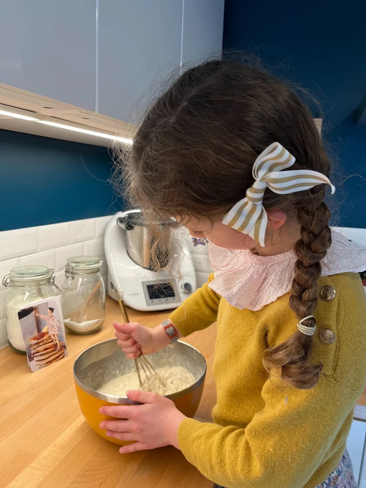 Petite fille préparant des crêpes, pull moutarde et nœud rayé doré — By Mésange