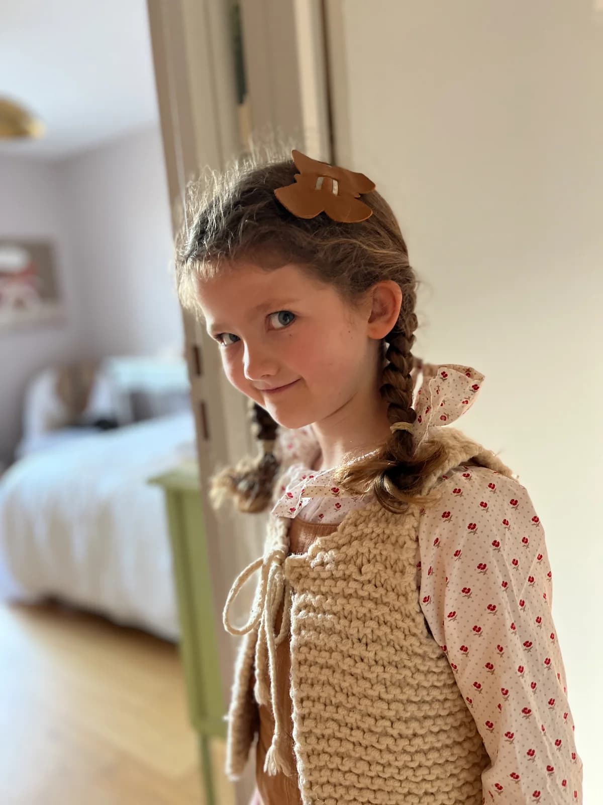 Fillette souriante en gilet tricot beige et blouse fleurie à smocks, barrette papillon — By Mésange