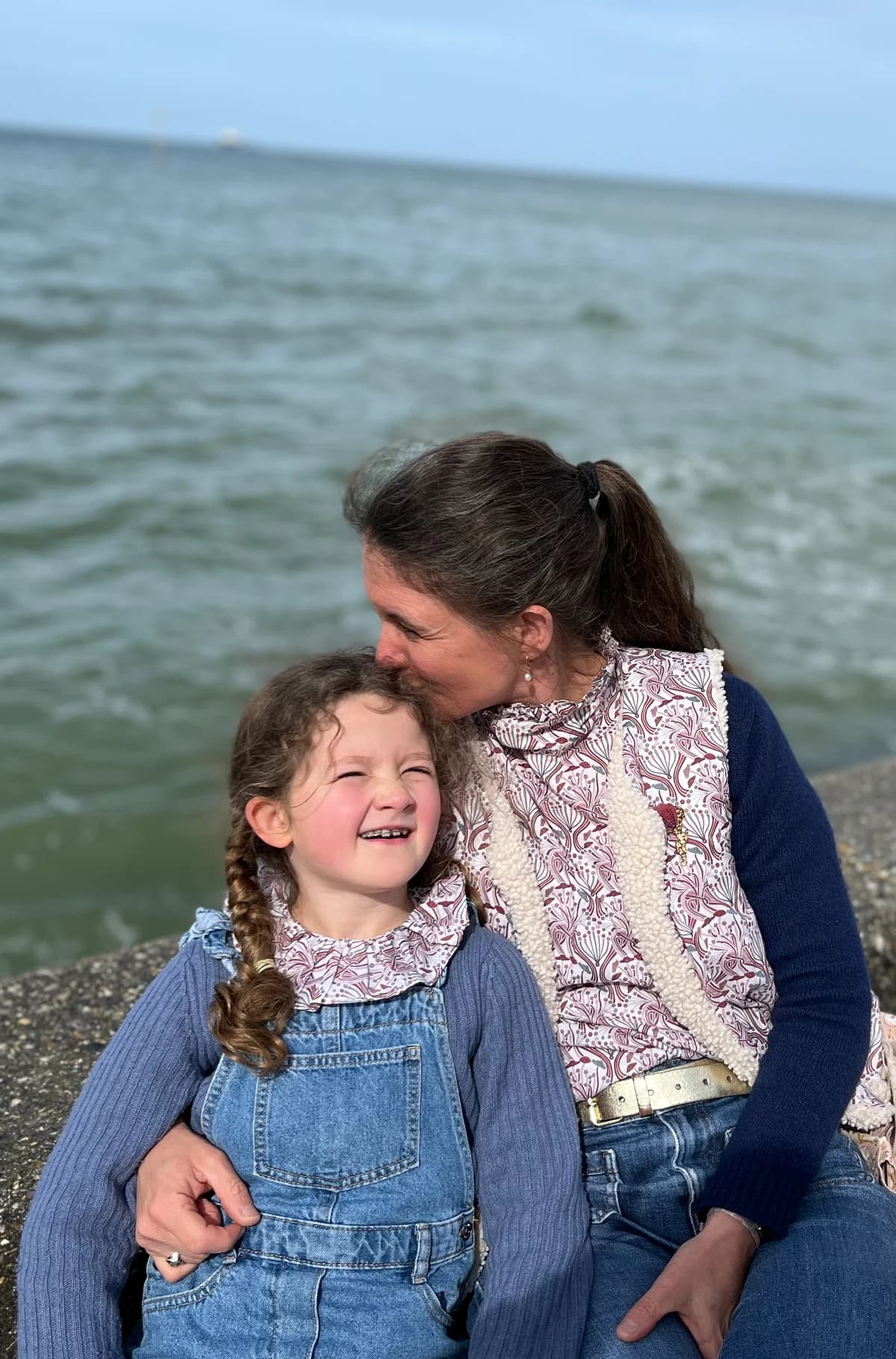 Stéphanie et sa fille assises au bord de mer, blouse paon et salopette en jean — By Mésange